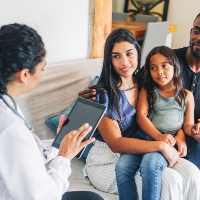 Young family sitting on sofa listening to the advice from a female doctor using digital tablet. Doctor on house call giving medical consultation to a young family.